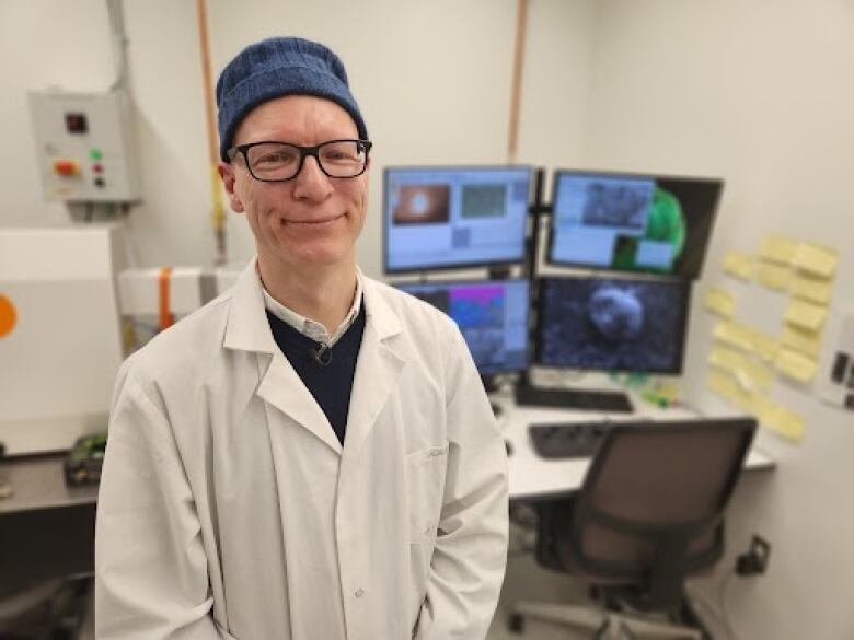 A man in glasses and a lab coat smiles at the camera in front of multiple screens showing microscopic images.