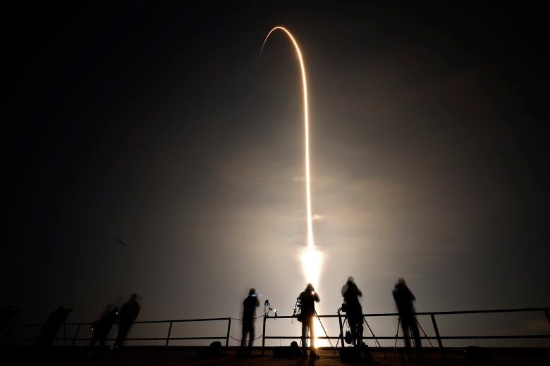 Onlookers and photographers are shown in the foreground, with the vapour trail in the night sky shown above them.