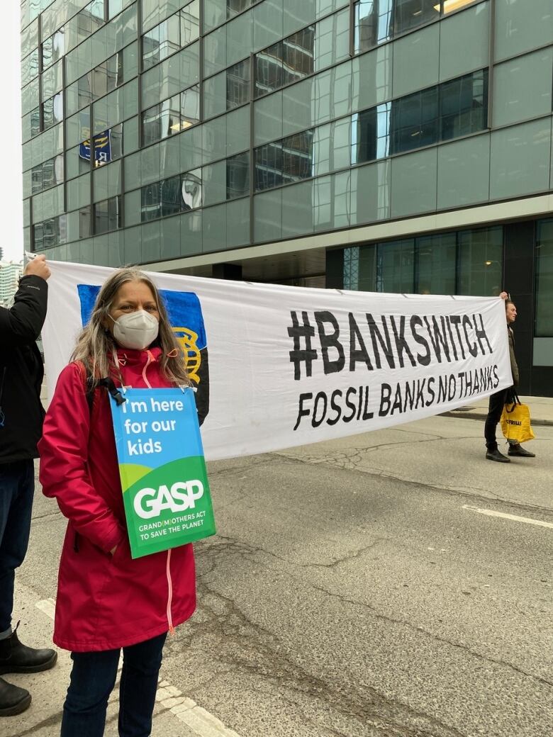 A woman with long hair and a red jacket stands on a street with a sign around her neck advocating for environmental sustainability.