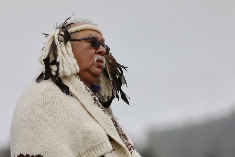 Eric Pelkey is shown in a close up shot standing outside against a grey sky. He is a middle-aged Indigenous man wearing a traditionally woven black and white sweater done in the Cowichan-style and a headdress of feathers and wool.