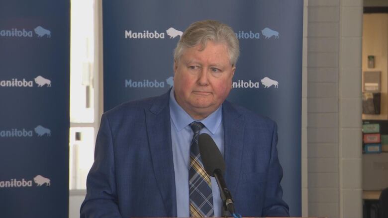 A man in a blue shirt and blue blazer speaks into a microphone at a podium.