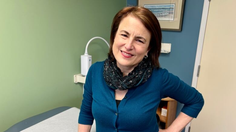 A woman in a doctor's office wearing a cardigan smiles at the camera.