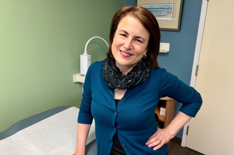 A woman in a doctor's office wearing a cardigan smiles at the camera.