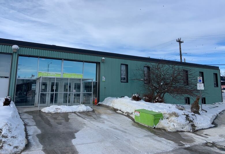 Snow surrounds a green building. A green sign above the door says 