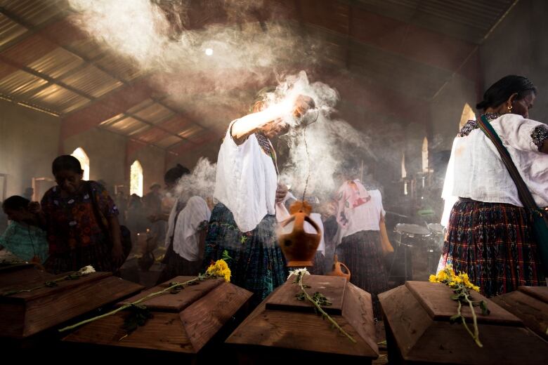 People stand in a dark, shaded room before a row of small coffins containing recovered remains.