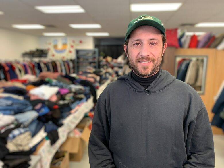 A man with a beard and ball cap in a gray hoodie stands between tables of clothes in the Free Store for Ukrainian Newcomers.