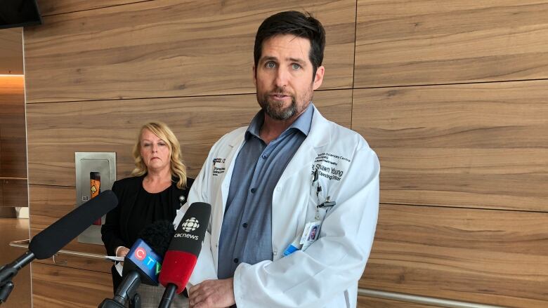 A woman with long blonde hair in a black top stands in the background as a man in a blue shirt and a lab coat with dark hair and a goatee stands behind three microphones.