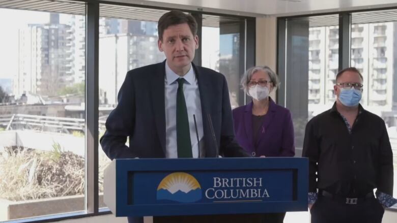 A man stands at the podium speaking, with two people standing behind him in front of glass windows.