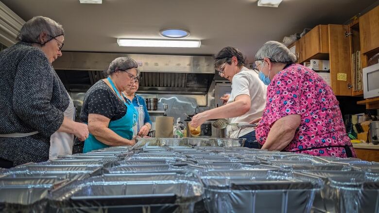 Five women in hair nets stand over a table full of tinfoil takeout containers. One woman pours a measuring cup full of beans into one container.