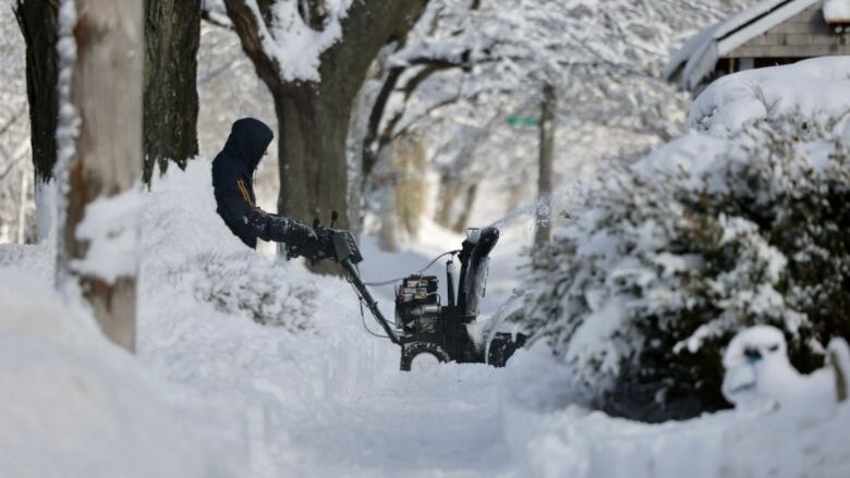 A man with his hood pulled up is seen operating a snow blower on his driveway from a sidewalk cleared of snow.