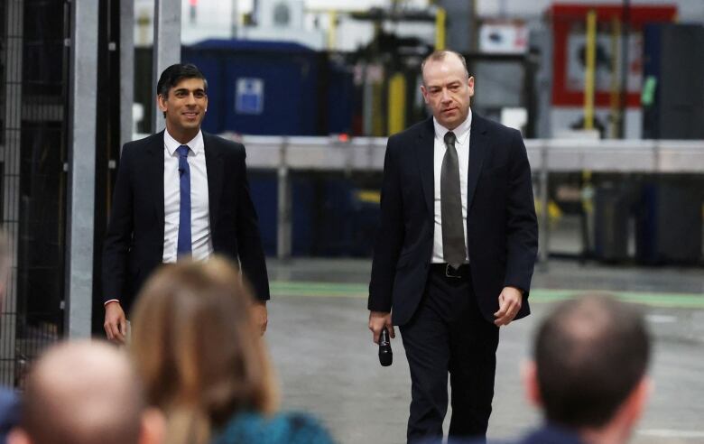 Prime Minister Rishi Sunak  and Northern Ireland Secretary Chris Heaton-Harris dressed in suits and walking side-by-side in a factory.