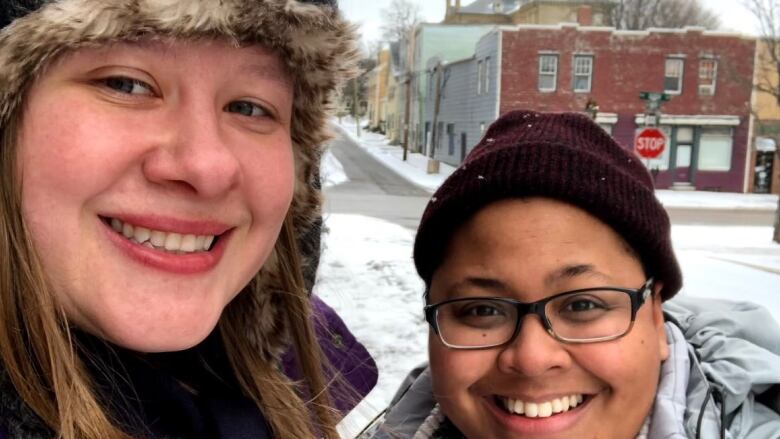 Two women in winter coats stand together in front of a snowy street.