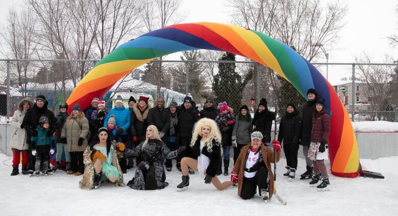 Children, parents, city officials and drag queens stand and smile for a photo underneath a rainbow archway on an outdoor rink.