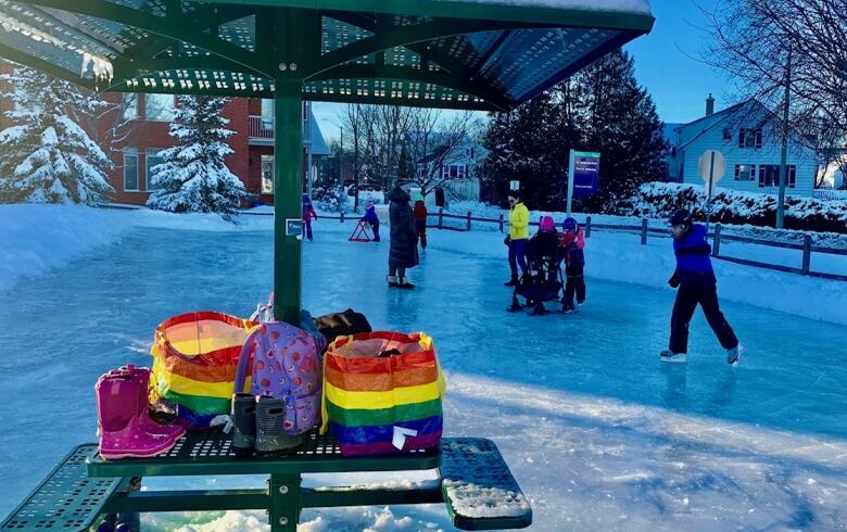 Children skate on a puddle rink in a small park. On the bottom right hand corner there is a rainbow-coloured bag on a park bench.