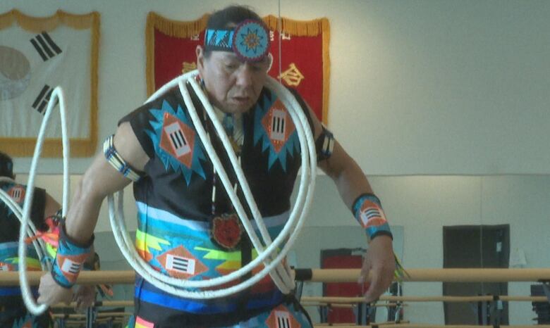 Wearing regalia coloured black, turquoise and orange, Pipestem prepares to roll a hoop on the ground. 