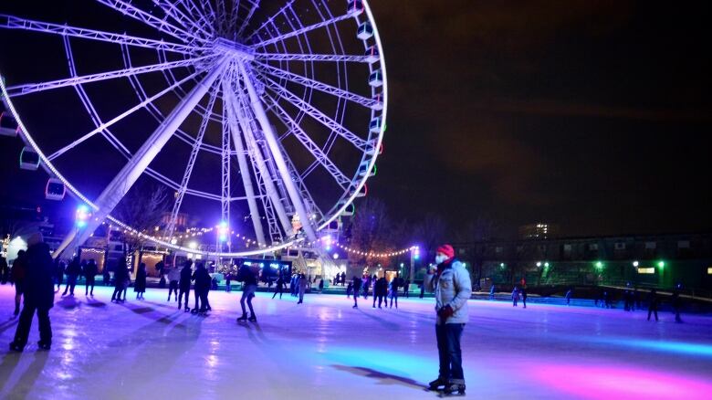 People skate at night with the coloured lights of a huge ferris wheel in the background.