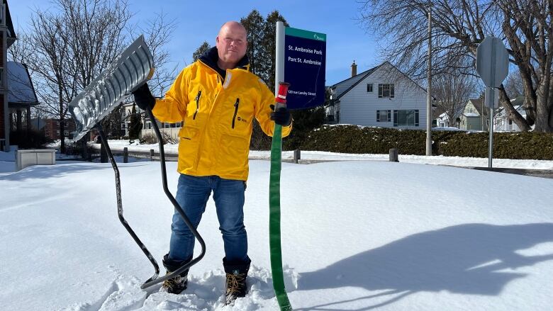 A man wearing a yellow jacket holds up a snow shovel and hose. Behind him is a sign for St. Ambroise Park in Vanier