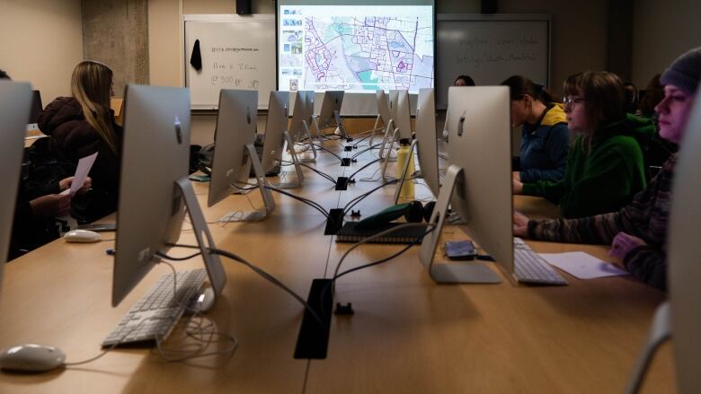 Students sat working on computers in a classroom with a map projected on a screen in the background.