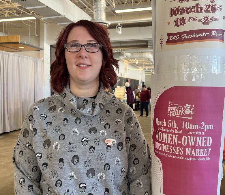 A young woman smiles for the camera. She stands next to a poster that's taped on a pillar and reads 