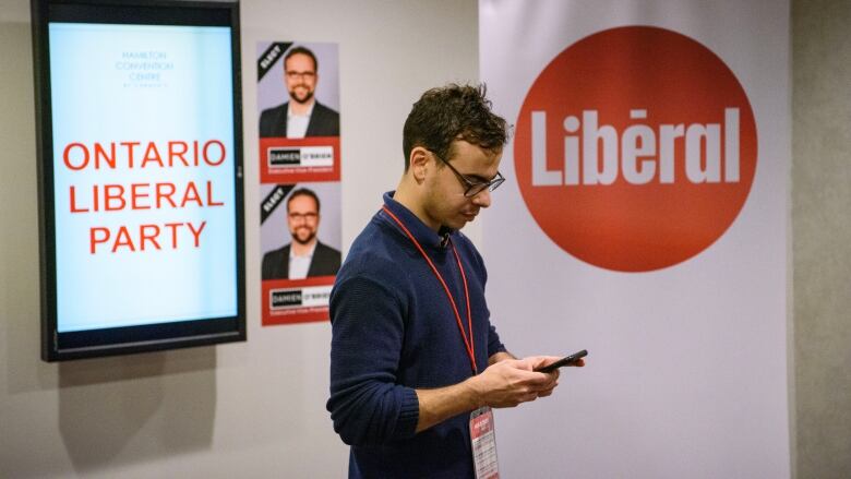 A man stares down at his phone. Behind him are signs for the Ontario Liberal Party.