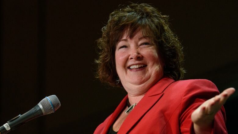 A woman is standing behind a microphone, smiling and gesturing with her open hand.