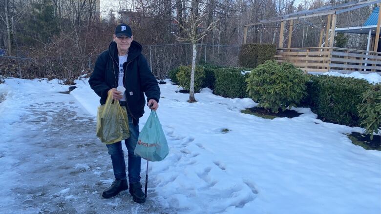 A man is pictured standing on a snowy sidewal, carrying two grocery bags. 