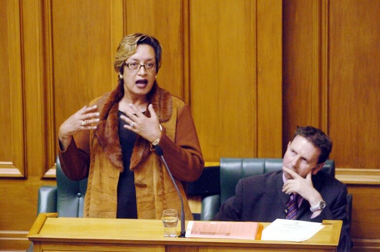 A person gestures with their hands while speaking inside a wood-paneled room, with a man shown watching and listening to them.