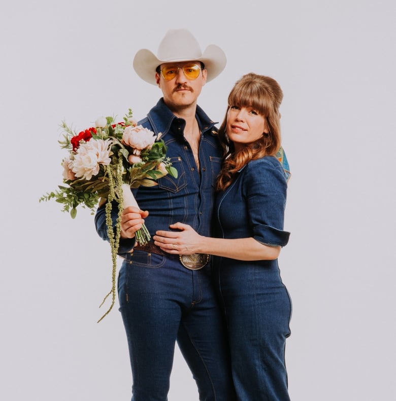 A married couple stands in denim outfits. The husband is holding a bouquet of flowers.