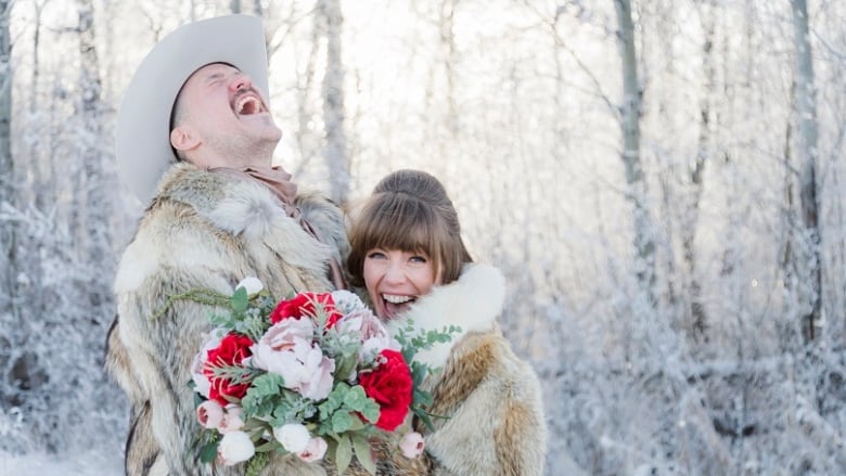 A smiling couple stands outside on a cold winter day wrapped in fur jackets. 