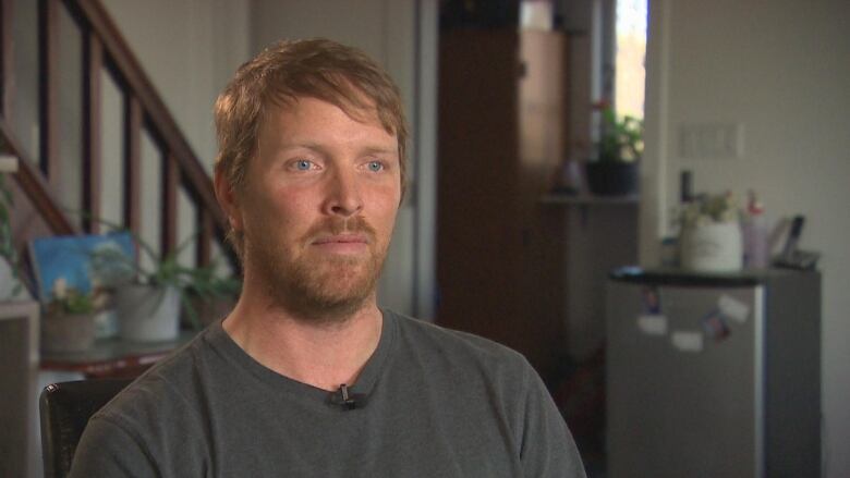 A portrait of a man with light brown hair and blue eyes wearing a dark grey t-shirt. 