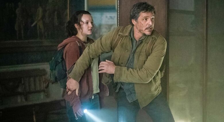 Still frame from the tv series The Last Of Us. Pedro Pascal steps in front of Bella Ramsey, holding her arm in a protective gesture. Pedro wears a khaki green work jacket and Bella wears a pink hoodie.