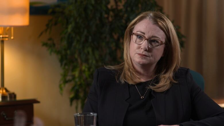 A woman wearing a black shirt and glasses looks towards the camera.