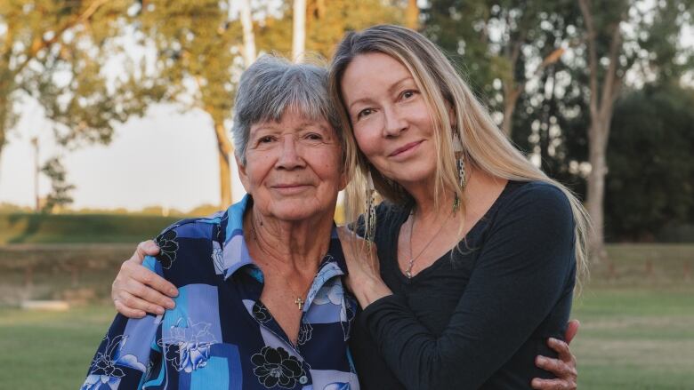 A younger woman and an older woman with a strong resemblance embrace while looking at the camera in a green area with trees behind them.