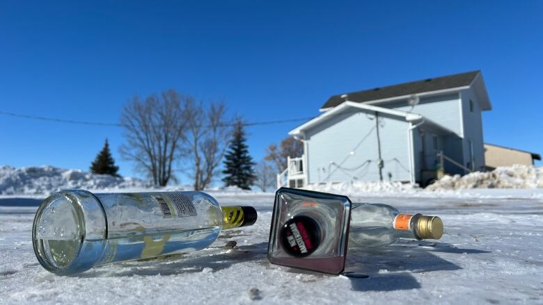 Three alcohol bottles are seen on a snowy ground with a house and trees in the background.