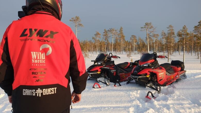 A in a red snowmobile jacket turned back on to the camera with two Bombardier Lynx snowmobiles in the background.