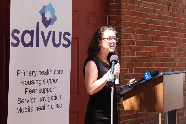 A woman wearing a dark coloured sleeveless shirt standing at a podium with several microphones in front of a red brick wall and a sign saying 