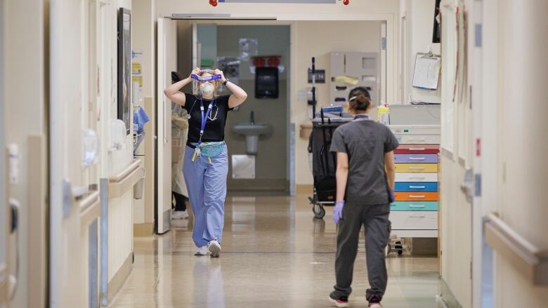 Two health care professionals walk down the empty hall of a hospital, putting on their personal protective equipment.