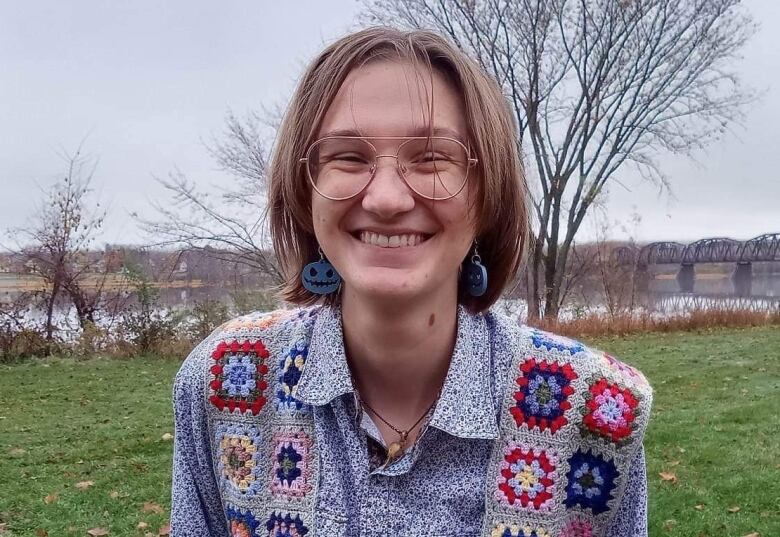 A person standing outside in a field with water in the background. They have chin length hair, glasses and a are wearing a crocheted vest.