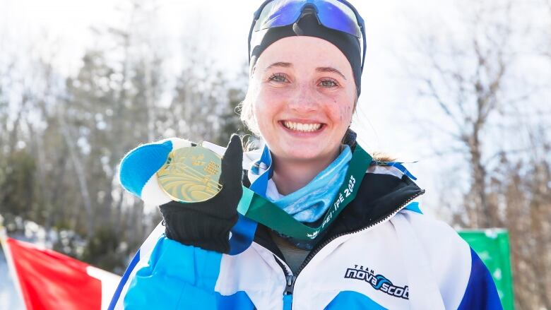 A young woman wearing ski gear holds a gold medal while smiling.