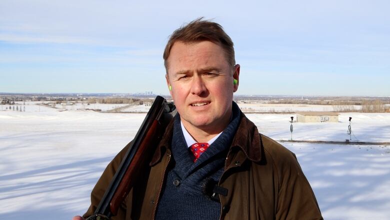 Man standing in a field with snow, carrying a gun on shoulder and wearing earplugs. 