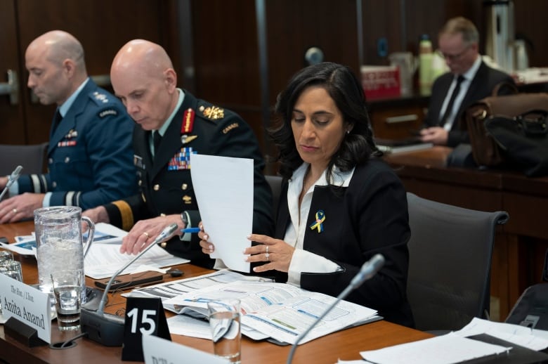 A woman sits at a table before a microphone holding a piece of paper, beside two men in military officer jackets.