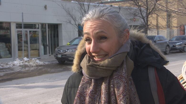 A woman with silver hair, wearing a coat and scarf is pictured standing in front of a street lined with cars.