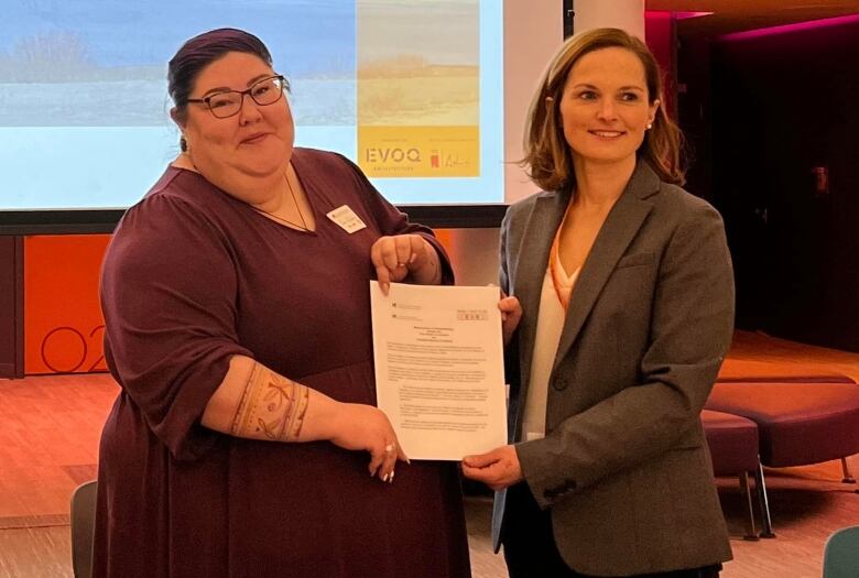 Two women stand holding a piece of paper and smiling at the camera. 