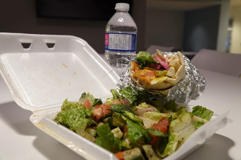 A salad covered in dressing is shown in a white Styrofoam container with a bottle of water in the background and a wrap with pickles, turnip and chicken sticking out of it. 