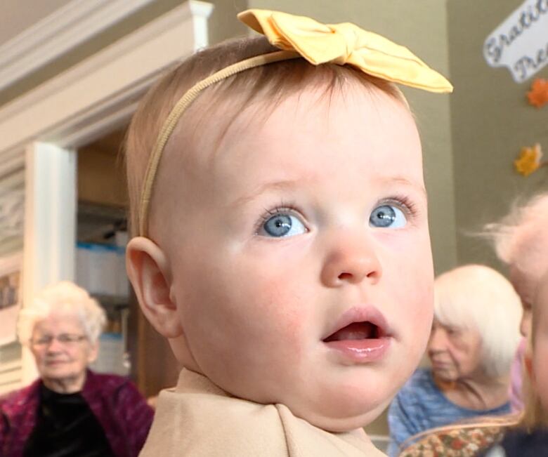 A baby girl wears a bow on a headband sits in the middle of a room. Seniors sit on couches on the edges of the room and look at her.