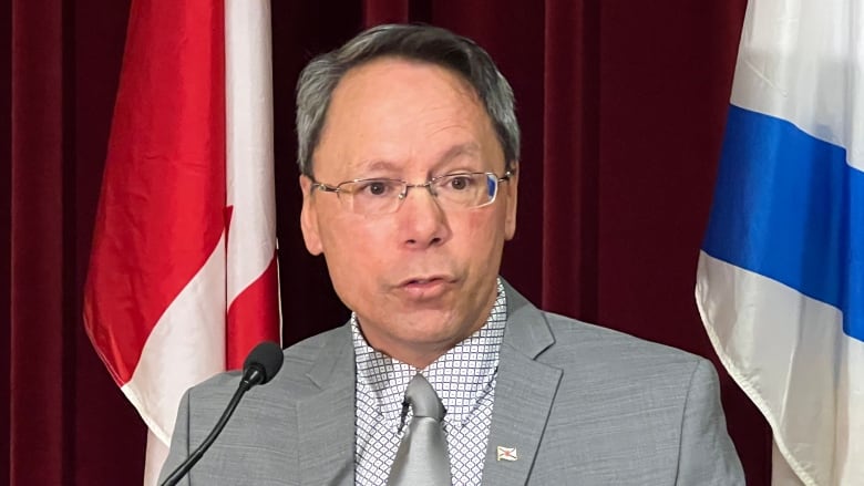 A man with a white dress shirt, grey jacket and grey tie stands in front of a Nova Scotia flag and a Canada flag.