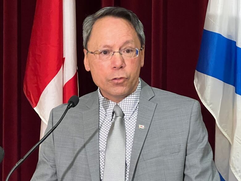 A man with a white dress shirt, grey jacket and grey tie stands in front of a Nova Scotia flag and a Canada flag.