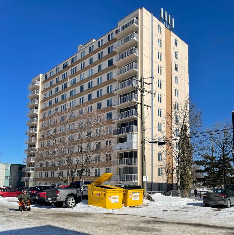 A highrise apartment building shot from the back shows yellow dumpsters, parked cars and a person in a mobility scooter.