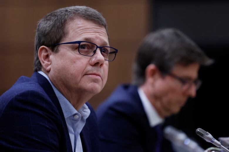 A man with brown hair, glasses and wearing a suit sits at a small microphone, next to another man in the background, and looks up.