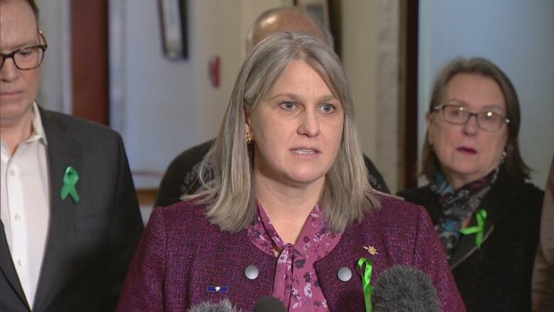 A woman with a green ribbon in her lapel stands flanked by a man and a woman, also wearing green ribbons, at a news conference.